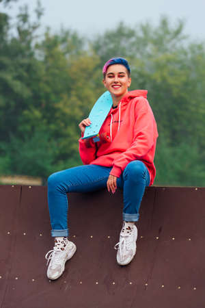 Young girl with short multicolored hair and nose piercing dressed in pink hoodie sitting with skateboard in skatepark during morning fog. Portrait of a tomboy with plastic skateboardの写真素材