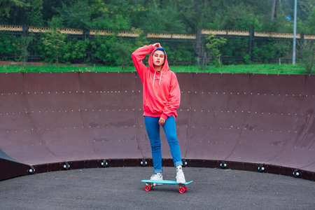 Portrait of a trendy pretty young girl dressed in pink hoodie and jeans standing next to the skateboard coart with her blue plastic skateboard.の写真素材