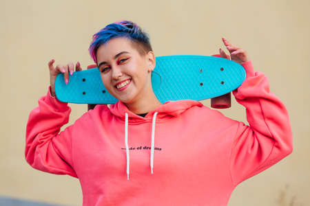 Stylish young woman with short colored hair holding blue plastic skateboard on her shoulders. Youth...
