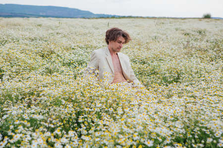 Tall handsome man dressed in a white suit on naked body sitting on a white chair in camomile flowers fieldの写真素材