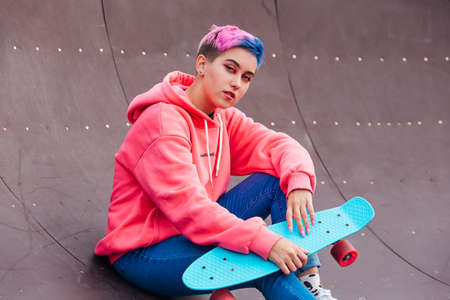 Stylish young woman with short colored hair sitting with her blue plastic skateboard in skatepark....