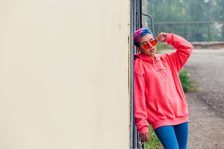 Young girl with short colorful hair and nose piercing dressed in pink hoodie and pink sunglasses...