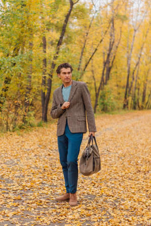Tall handsome man dressed in a brown jacket walking with a bag on the autumn alleyの写真素材