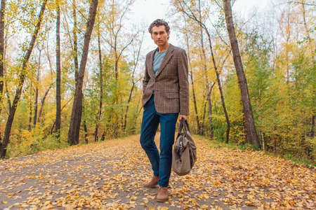 Tall handsome man dressed in a brown jacket walking with a bag on the autumn alleyの写真素材