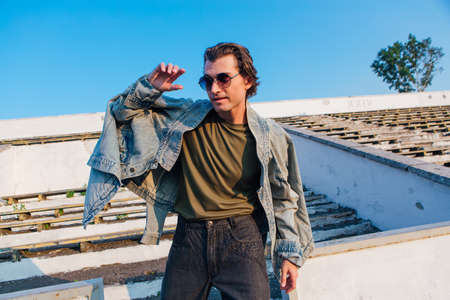 Fashion portrait of a tall handsome man dressed in jeans jacket ans sunglasses on the crushed tribune of the old stadium during sunsetの写真素材
