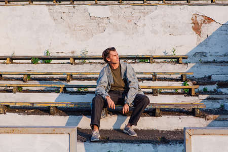 Fashion portrait of a tall handsome man dressed in jeans jacket on the crushed tribune of the old stadium during sunsetの写真素材