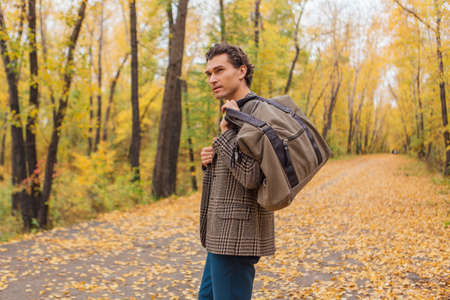 Tall handsome man dressed in a brown jacket walking with a bag on the autumn alley