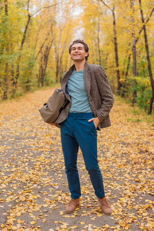 Tall handsome man dressed in a brown jacket walking with a bag on the autumn alleyの写真素材