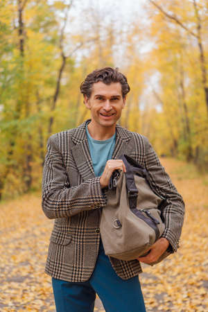 Tall handsome man dressed in a brown jacket walking with a bag on the autumn alley