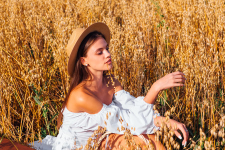 Rural Countryside Scene. Young beautiful woman with long hair dressed in white blouse and straw hat sitting at golden oat field near the basket with ears of oats.の写真素材