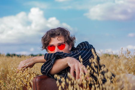 Rural Countryside Scene. Tall handsome man dressed in a black shirt and pink sunglasses laying at golden oat field with brown vintage leather suitcase full of ears of oats. Summer landscape with blue skyの写真素材