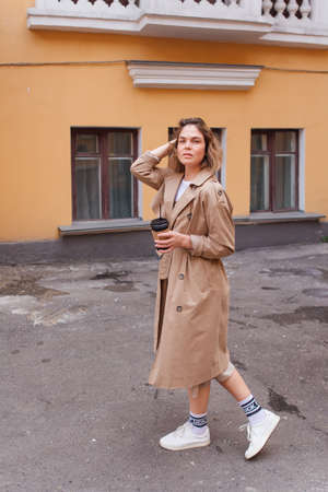 Young smiling millennial woman with wild hair dressed in an autumn coat walking with a cup of coffee to go near the old building.の写真素材