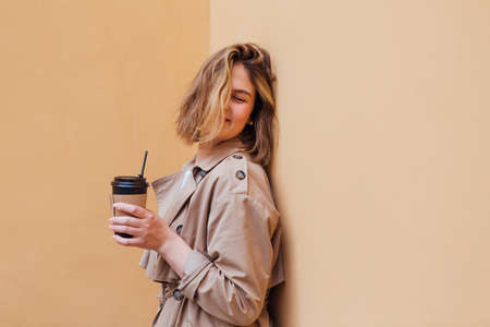 Young smiling millennial woman with wild hair dressed in an autumn coat standing a cup of coffee to...