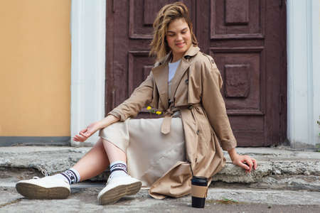 Young smiling millennial woman with wild hair dressed in an autumn coat sitting near the door of an old building with a cup of coffee to go.の写真素材