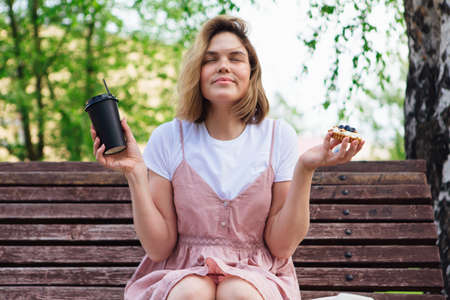 Young romantic woman sitting on bench in park with cake and cup of coffee to go in a sunny summer...
