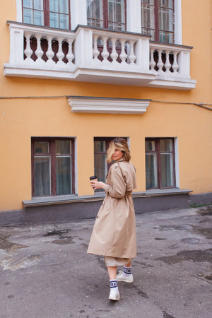 Young millennial woman with wild hair dressed in an autumn coat walking with a cup of coffee to go near the old building.の写真素材