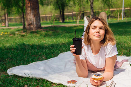 Young romantic woman laying on the grass in park with open book, cake and cup of coffee to go in a sunny summer dayの写真素材