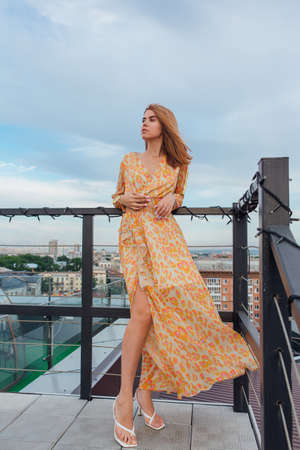 Beautiful young woman in long leopard dress standing on the stairs on the top of the roof of a building.の写真素材