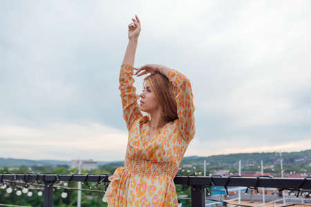Beautiful young woman in long leopard dress standing on the stairs on the top of the roof of a building.の写真素材