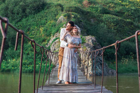 Beautiful young romantic couple standing and hugging on the suspension bridge above the riverの写真素材