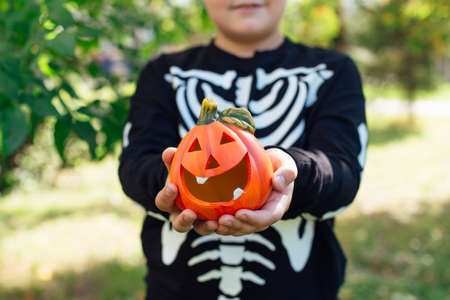 Smiling boy in black costume of skeleton holding small decorative halloween pumpkin outdoors.の写真素材