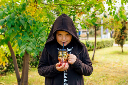 Smiling boy in black costume of skeleton and hood holding glass in the shape of a skull outdoors.の写真素材