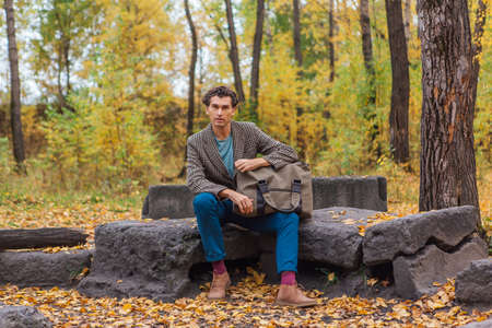 Tall handsome man with a bag sitting on the rocks in the autumn forestの写真素材