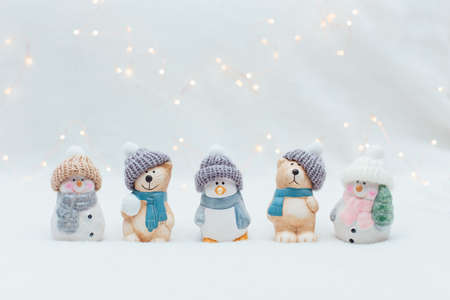 Decorative Christmas-themed figurines. The statuette of a cat, a penguin, a bear and two snowmen in a knitted hat on the white background. Festive decor, warm bokeh lightsの写真素材