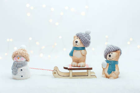 Decorative Christmas-themed figurines. The statuette of a cat, snowman and a bear on the sledge. Cute animals in a knitted hat on the white background. Festive decor, warm bokeh lightsの写真素材