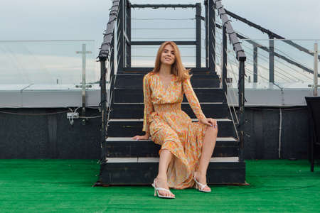 Beautiful young woman in long leopard dress sitting with bare feet on the stairs on the top of the roof of a building.の写真素材