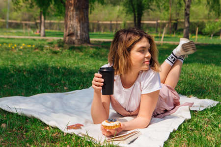 Young romantic woman laying on the grass in park with open book, cake and cup of coffee to go in a sunny summer dayの写真素材