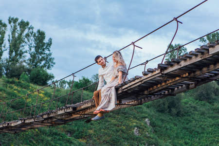 Beautiful young romantic couple sitting and hugging on the suspension bridge above the riverの写真素材