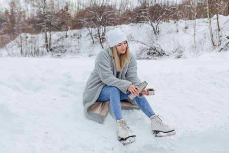 Lovely young woman relaxing after riding ice skates and drinking hot drink from termo pot on the ice rink. Girl sitting on snow and smiling.の写真素材