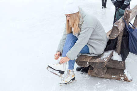 Lovely young woman sitting on the bench and putting on figure skates on the ice rink. Girl is going to skate on ice in a winter frosty dayの写真素材