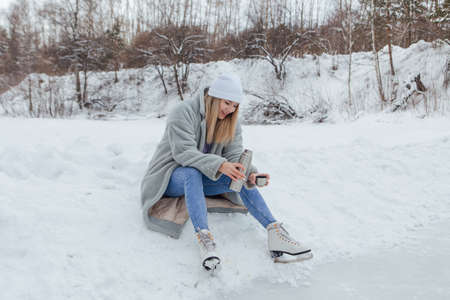 Lovely young woman relaxing after riding ice skates and drinking hot drink from termo pot on the ice rink. Girl sitting on snow and smiling.の写真素材