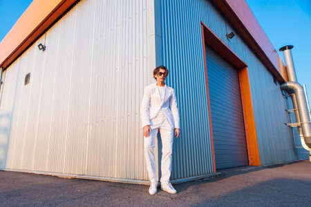 Wide angle portrait of a handsome man dressed in a white suit and round hippie sunglasses standing near the corner of a building during sunsetの写真素材