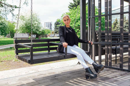 Young stylish business woman with short hair and nose piercing. Confident girl look like lesbian sitting with mobile phone on the swingの写真素材