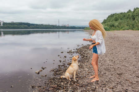 Young beautiful woman with blond curly hair trains her labrador retriever dog on the river shoreの写真素材