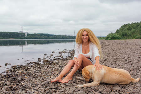 Young beautiful woman with blond curly hair sitting with her labrador retriever dog on the river shoreの写真素材