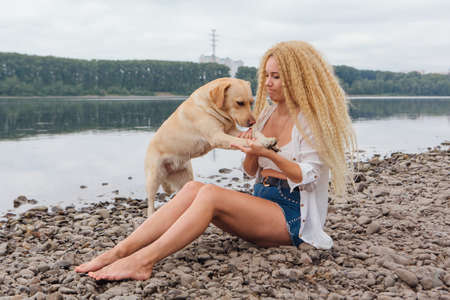 Young beautiful woman with blond curly hair sitting with her labrador retriever dog on the river shoreの写真素材