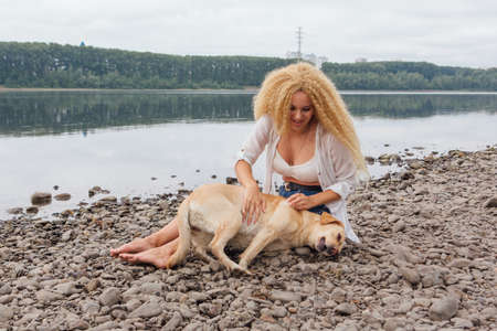Young beautiful woman with blond curly hair sitting with her labrador retriever dog on the river shoreの写真素材