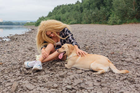 Young beautiful woman with blond curly hair sitting with her labrador retriever dog on the river shoreの写真素材
