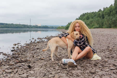 Young beautiful woman with blond curly hair sitting with her labrador retriever dog on the river shoreの写真素材