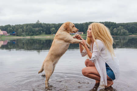 Young beautiful woman with blond curly hair huging with her labrador retriever dog on the river shoreの写真素材