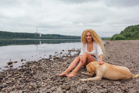 Young beautiful woman with blond curly hair sitting with her labrador retriever dog on the river shoreの写真素材