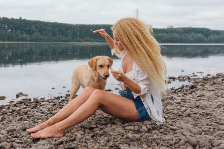 Young beautiful woman with blond curly hair sitting with her labrador retriever dog on the river shoreの写真素材