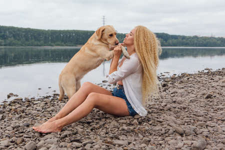 Young beautiful woman with blond curly hair sitting with her labrador retriever dog on the river shoreの写真素材