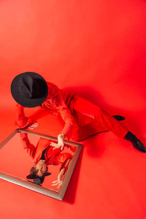 Tall handsome man dressed in red shirt and black hat sitting on the red background and looking into the mirror on his own reflection.の写真素材