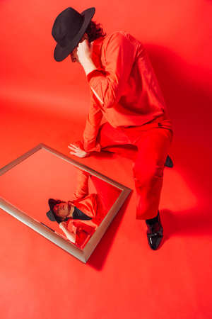 Tall handsome man dressed in red shirt and black hat sitting on the red background and looking into the mirror on his own reflection.の写真素材