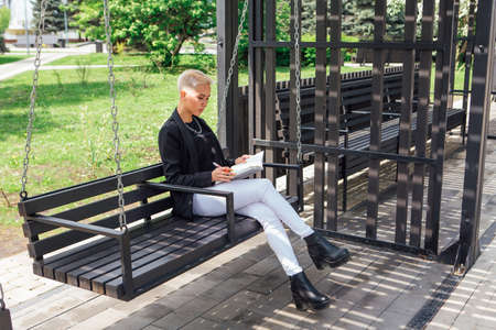 Young stylish business woman with short hair and nose piercing. Confident girl look like lesbian sitting with notepad on the swingの写真素材